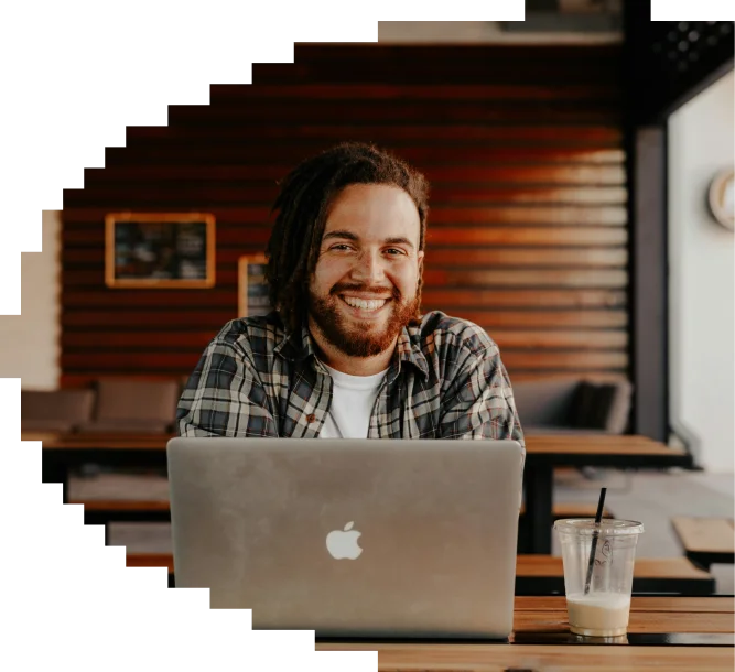 Smiling man with dreadlocks wearing a plaid shirt, using a MacBook at an outdoor café with a drink nearby.