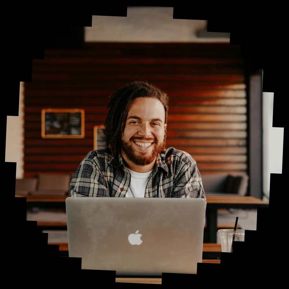 Smiling man wearing a plaid shirt, using a MacBook laptop in a cozy café setting.