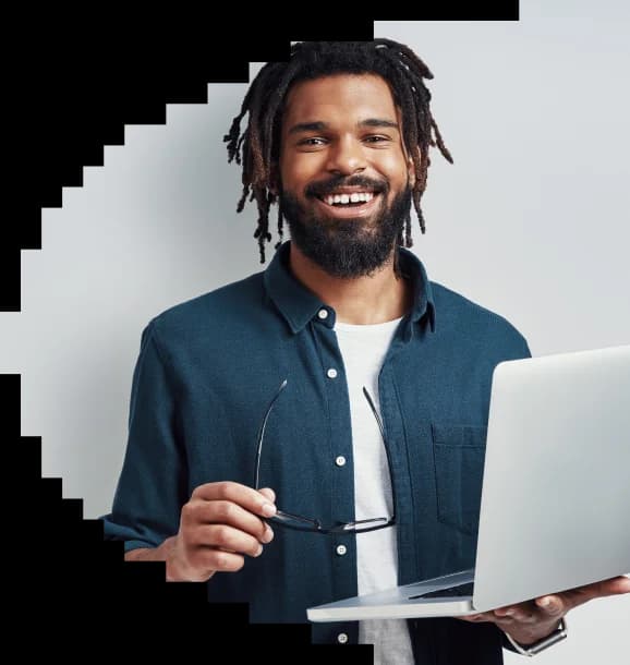 Smiling man with dreadlocks wearing a dark blue shirt, holding glasses and using a laptop against a white wall.