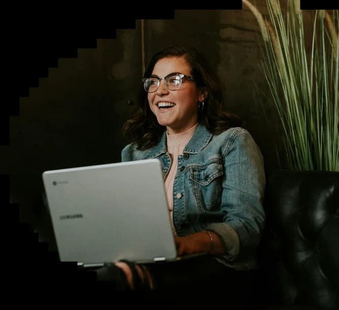 Smiling woman in glasses and a denim jacket using a Samsung laptop while sitting on a black couch.